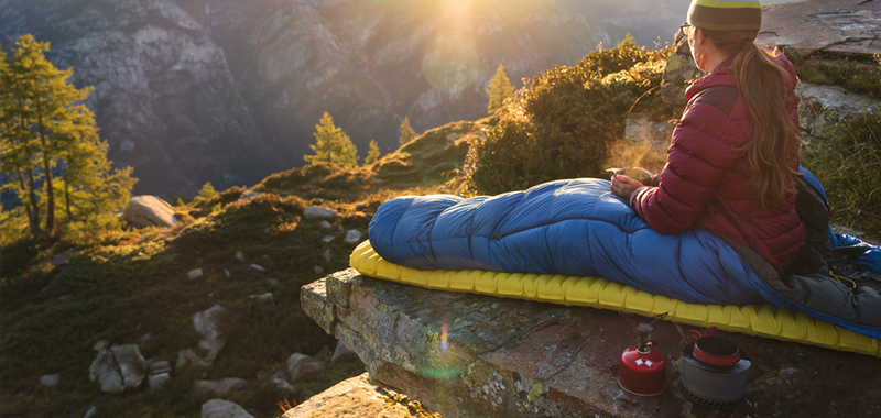 Frau mit Schlafsack sitzt auf Isomatte in der Natur